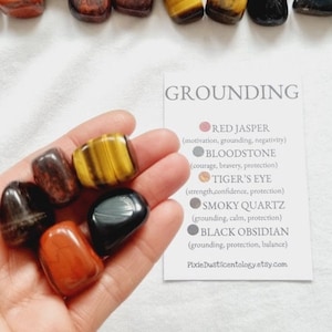 Peut inclure: Une collection de cristaux d'ancrage, y compris le jaspe rouge, la pierre de sang, l'œil de tigre, le quartz fumé et l'obsidienne noire. Chaque cristal est étiqueté avec son nom et ses propriétés. Le texte sur la carte indique "GROUNDING" et liste les propriétés de chaque cristal. L'adresse du site Web est PixieDustScentology.etsy.com.