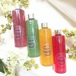 Puede incluir: Cuatro botellas transparentes llenas de l&iacute;quido colorido y texturizado. Las botellas est&aacute;n etiquetadas como "Lavanda", "Aloe Vera", "Vitamina C" y "Rosa". Cada botella tiene una tapa plateada. El fondo incluye flores blancas y una tela blanca.