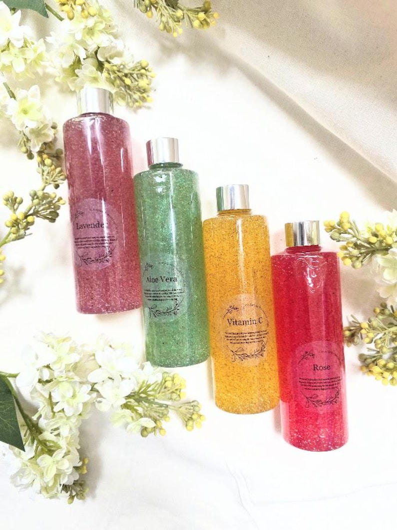Puede incluir: Cuatro botellas transparentes llenas de l&iacute;quido colorido y texturizado. Las botellas est&aacute;n etiquetadas como "Lavanda", "Aloe Vera", "Vitamina C" y "Rosa". Cada botella tiene una tapa plateada. El fondo incluye flores blancas y una tela blanca.