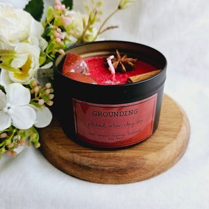 May include: A black candle tin with a red soy wax candle inside. The candle is infused with red jasper crystals and has a grounding scent of patchouli, vetiver, and clary sage.