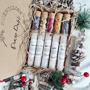 May include: A set of six glass test tubes filled with bath salts and dried flowers. The tubes are labeled with different scents: "Unwind", "Away", "Self-Love", "Uplift", "Relax". The tubes are arranged in a brown cardboard box with shredded paper.