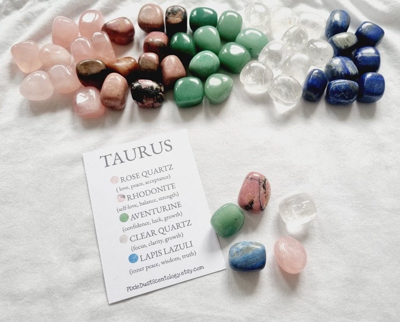 Op de afbeelding: Een set getrommelde stenen voor het sterrenbeeld Stier. De stenen zijn rozenkwarts, rhodonite, aventurijn, helder kwarts en lapis lazuli. De stenen zijn gerangschikt in een cirkel rond een kaart met de naam van het sterrenbeeld en de eigenschappen van elke steen.