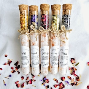 May include: Five glass test tubes filled with Himalayan salt and dried flowers. Each tube has a cork stopper and a label with a different name: Uplift, Insind, Self-Love, Anxiety, and Relax. The labels also list ingredients, including essential oils and dried flowers.