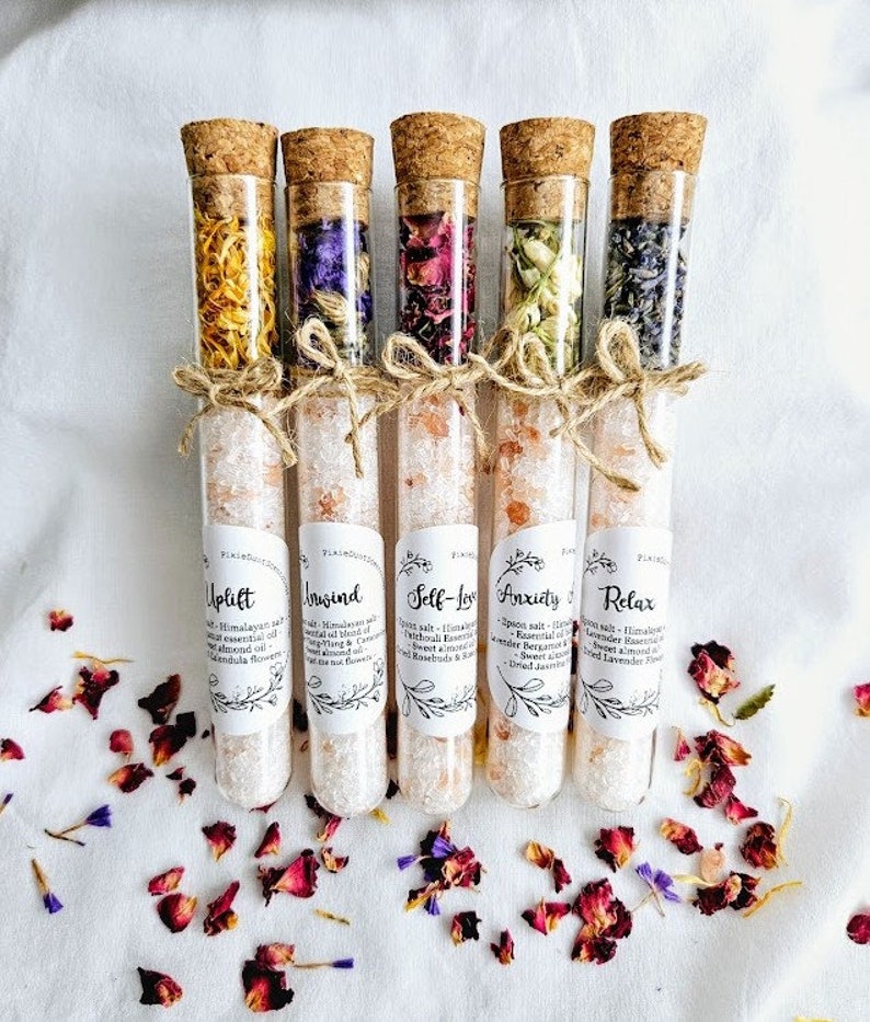 May include: Five glass test tubes filled with Himalayan salt and dried flowers. Each tube has a cork stopper and a label with a different name: Uplift, Insind, Self-Love, Anxiety, and Relax. The labels also list ingredients, including essential oils and dried flowers.