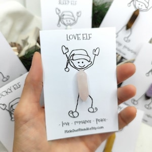 Peut inclure: Une carte blanche avec un lutin de dessin animé tenant une pointe de cristal rose. La carte indique "LOVE ELF - love - romance - peace - PixieDustBeads.etsy.com".