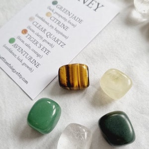 May include: A set of six tumbled stones for prosperity and abundance. The stones are green jade, citrine, clear quartz, tiger's eye, aventurine, and a white stone. The stones are arranged on a white surface with a card that lists the names of the stones and their properties.
