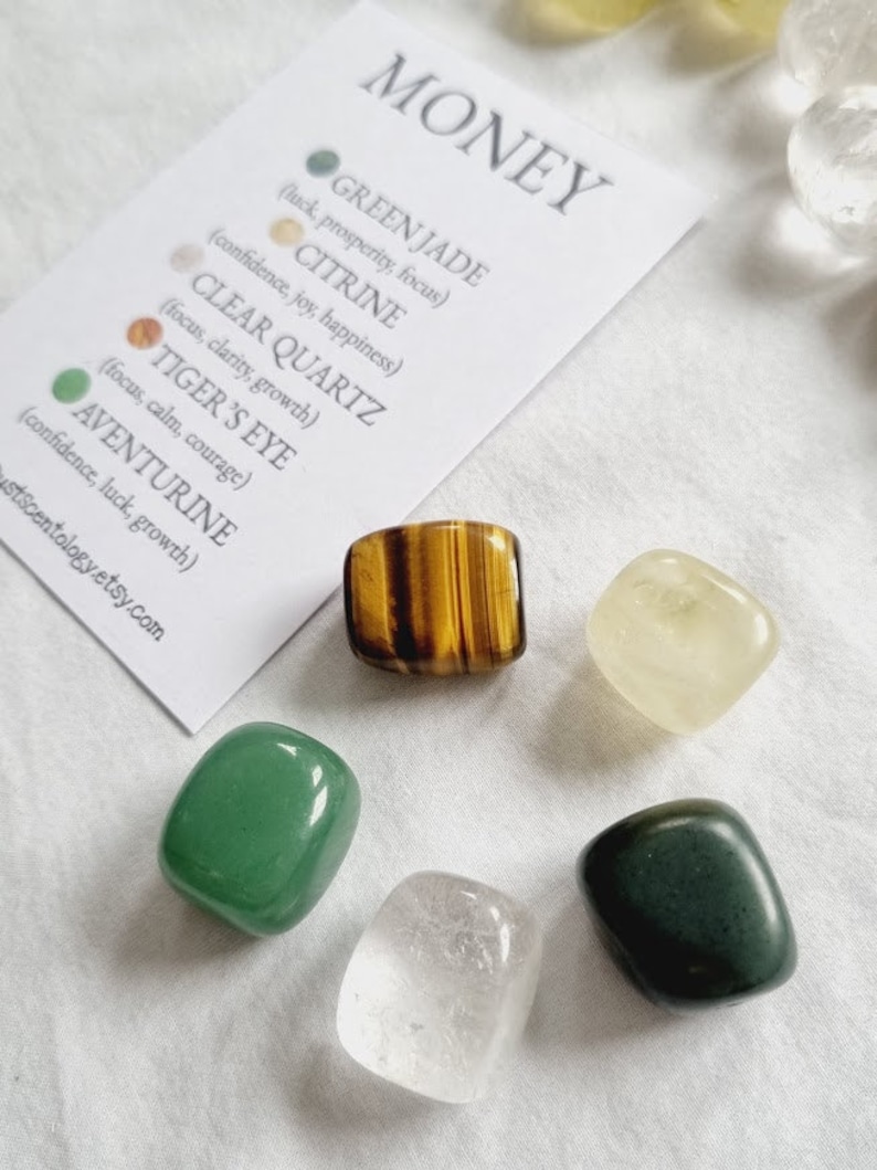 May include: A set of six tumbled stones for prosperity and abundance. The stones are green jade, citrine, clear quartz, tiger's eye, aventurine, and a white stone. The stones are arranged on a white surface with a card that lists the names of the stones and their properties.