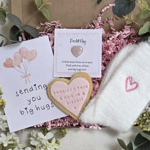 Op de afbeelding: Cadeaudoos met een wenskaart met ballonnen en de tekst "sending you big hugs", een hartvormige koek met de tekst "consider this a hug in a biscuit", een klein roze hart en witte fluffy sokken met een roze hart.