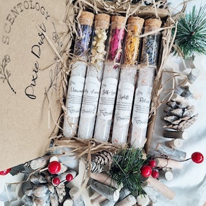 May include: A set of six glass test tubes filled with dried flowers and bath salts. The test tubes are labeled with names like "Unwind" and "Self-Love". The test tubes are arranged in a wooden box with a brown paper lid. The box is surrounded by pine branches and pine cones.