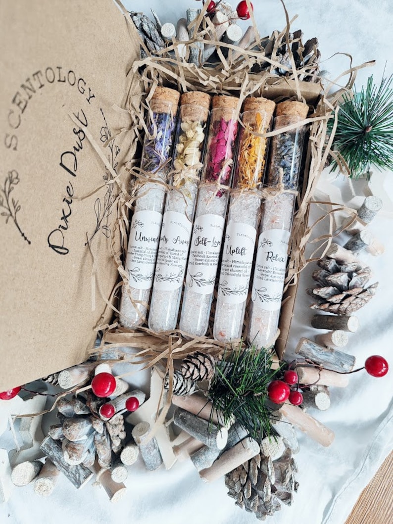 May include: A set of six glass test tubes filled with dried flowers and bath salts. The test tubes are labeled with names like "Unwind" and "Self-Love". The test tubes are arranged in a wooden box with a brown paper lid. The box is surrounded by pine branches and pine cones.