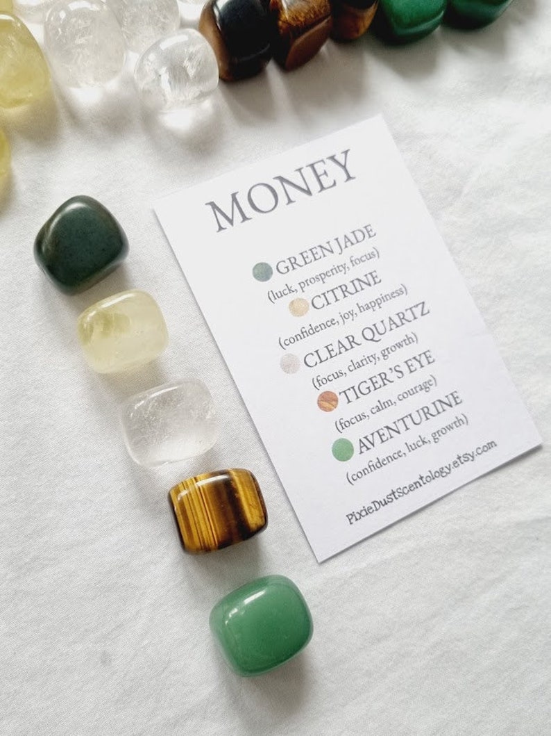 May include: A set of seven tumbled stones for money manifestation. The stones are green jade, citrine, clear quartz, tiger's eye, and aventurine. The stones are arranged on a white surface with a card that lists the stones and their properties.
