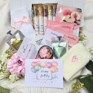 Könnte beinhalten: Eine Geschenkbox mit Pflegeprodukten. Sie enthält ein weißes Handtuch mit rosa Schleife, Badesalze in Glasröhrchen, Augenmasken, eine Karte mit "Happy Birthday", ein rosa Scrunchie und cremefarbene, flauschige Socken mit einem rosa Herz.