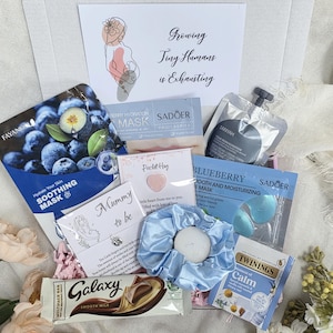 May include: A gift box filled with self-care items for a "Mummy to be". Includes face masks, a scrunchie, a chocolate bar, and tea. The box has a card with a drawing of a pregnant person and the text "Growing Tiny Humans is Exhausting".