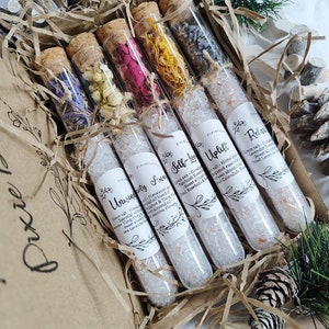 May include: Six glass test tubes filled with dried flowers and bath salts. The tubes are labeled with names like "Unwind" and "Uplift".