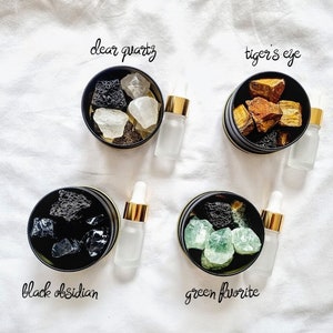 May include: Four small black containers with lids, each containing different types of crystals and a small glass bottle. The containers are labelled "clear quartz", "tiger's eye", "black obsidian", and "green fluorite".