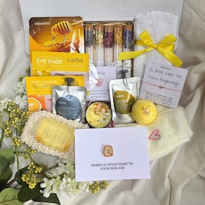 Pode incluir: Uma caixa de presente cheia de itens de autocuidado. Inclui máscaras para os olhos, bombas de banho, sais de banho, uma toalha pequena com um laço amarelo e um cartão com o texto "Here's a little toast to your new job".