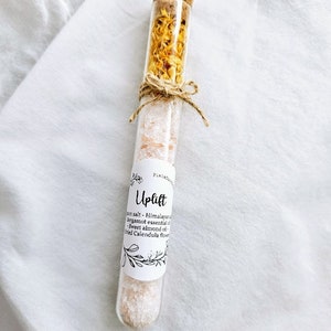 May include: A glass test tube filled with Himalayan salt, dried calendula flowers, and essential oils. The label reads "Uplift" and lists the ingredients: Himalayan salt, bergamot essential oil, sweet almond oil, and dried calendula flowers.
