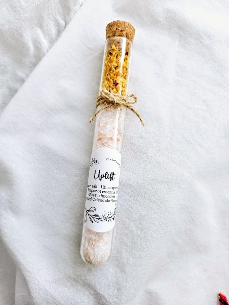 May include: A glass test tube filled with Himalayan salt, dried calendula flowers, and essential oils. The label reads "Uplift" and lists the ingredients: Himalayan salt, bergamot essential oil, sweet almond oil, and dried calendula flowers.