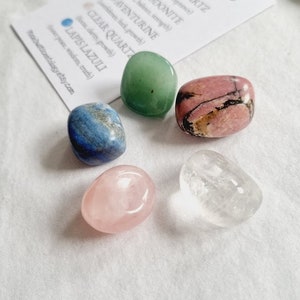 Op de afbeelding: Een set van vijf getrommelde stenen, waaronder rozenkwarts, rhodonite, aventurijn, helder kwarts en lapis lazuli. De stenen zijn gerangschikt op een wit oppervlak met een kaart waarop "Taurus" staat en de namen van de stenen en hun bijbehorende eigenschappen worden vermeld.