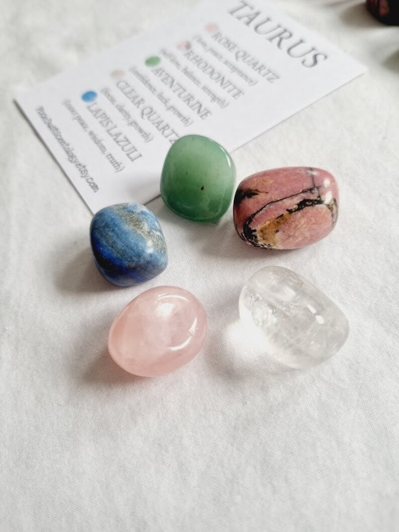 Op de afbeelding: Een set van vijf getrommelde stenen, waaronder rozenkwarts, rhodonite, aventurijn, helder kwarts en lapis lazuli. De stenen zijn gerangschikt op een wit oppervlak met een kaart waarop "Taurus" staat en de namen van de stenen en hun bijbehorende eigenschappen worden vermeld.