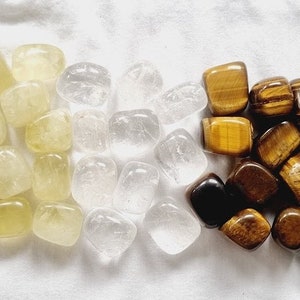 May include: A collection of tumbled stones in various colors, including green, yellow, white, brown, and black. The stones are arranged in a row on a white surface.