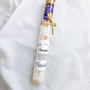 May include: A glass test tube filled with pink Himalayan salt, dried purple flowers, and a cork stopper. The label reads "Unwind" and lists ingredients: Himalayan salt, essential oil blend of Ylang-Ylang & Mandarin, sweet almond oil, and forget-me-not flowers.