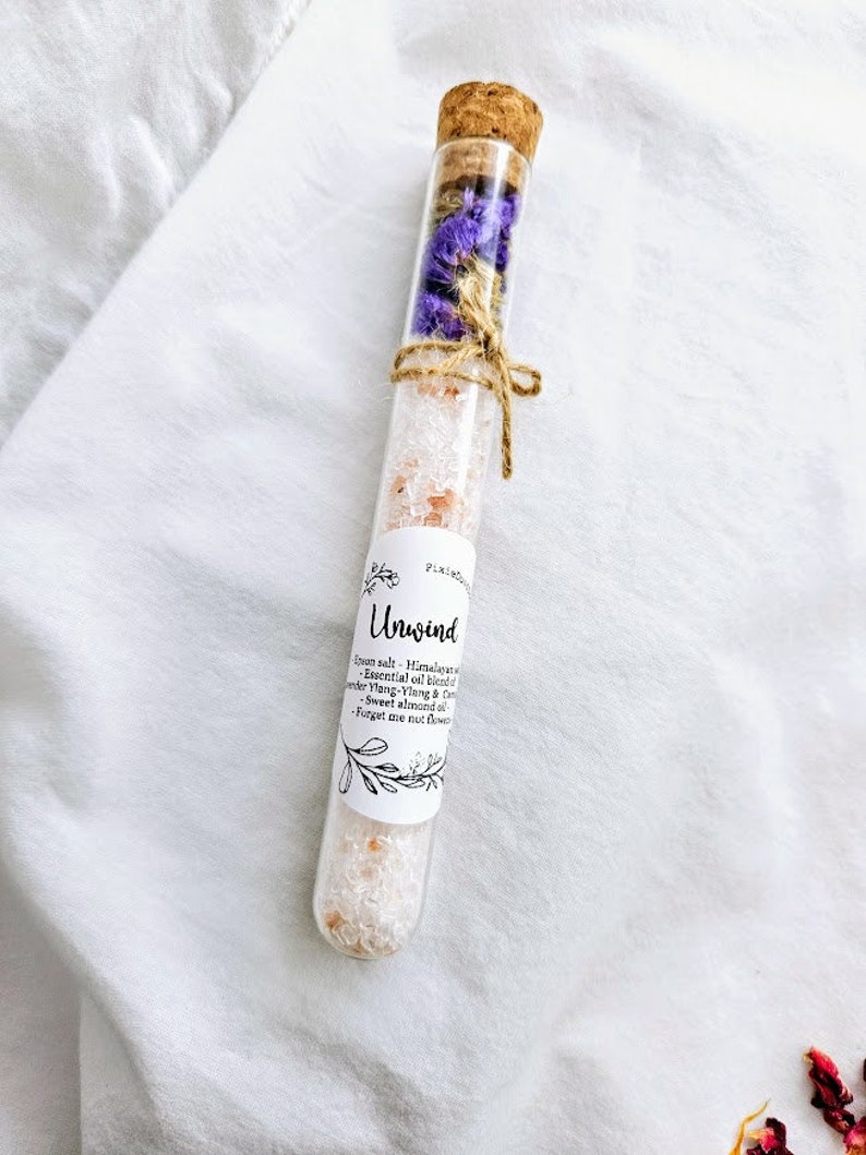 May include: A glass test tube filled with pink Himalayan salt, dried purple flowers, and a cork stopper. The label reads "Unwind" and lists ingredients: Himalayan salt, essential oil blend of Ylang-Ylang & Mandarin, sweet almond oil, and forget-me-not flowers.