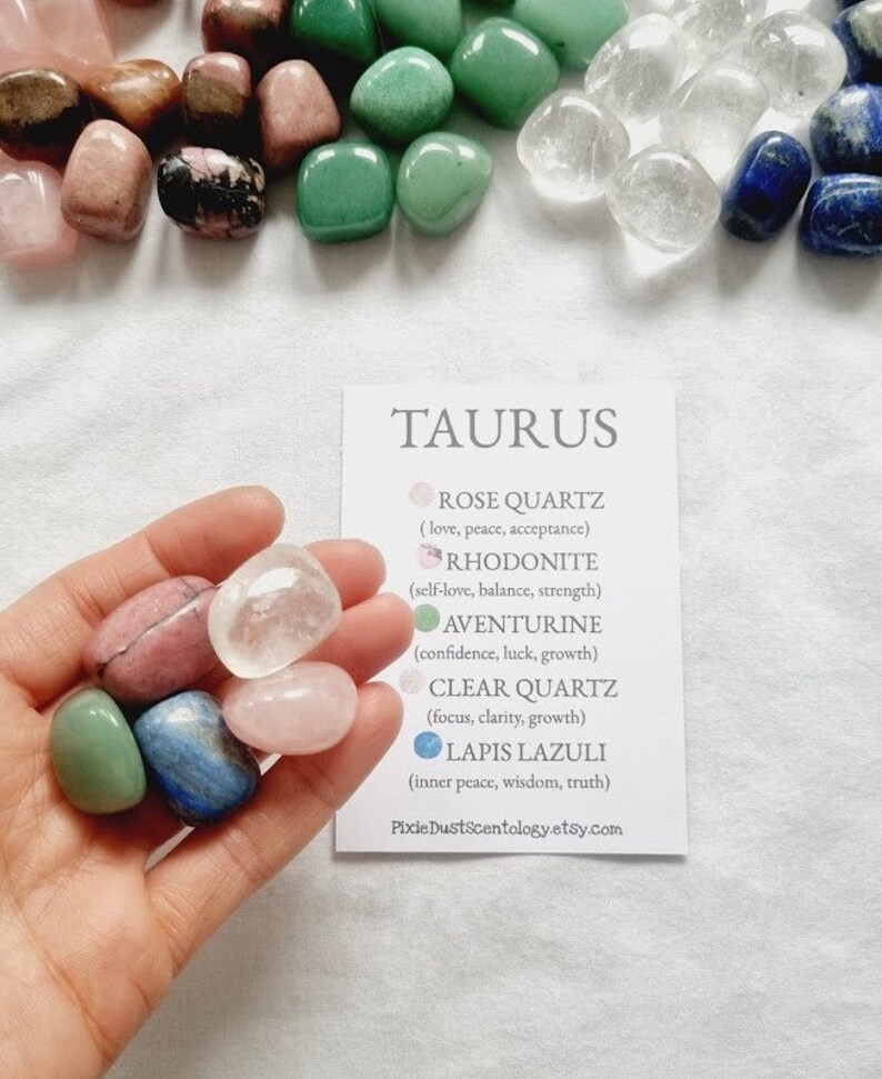 Op de afbeelding: Een set kristallen voor het sterrenbeeld Stier. De kristallen zijn rozenkwarts, rhodonite, aventurijn, helder kwarts en lapis lazuli. De kristallen zijn gerangschikt op een witte kaart met de naam van het sterrenbeeld en de eigenschappen van elk kristal.