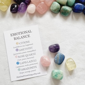 Könnte beinhalten: Ein Set an Trommelsteinen für emotionales Gleichgewicht, darunter Citrin, Amethyst, Rosenquarz, Lapislazuli und Amazonit. Jeder Stein ist mit seinem entsprechenden emotionalen Nutzen beschriftet. Die Steine sind auf einer weißen Oberfläche mit einer Karte angeordnet, auf der "Emotionales Gleichgewicht" steht.