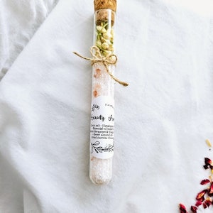 May include: A glass test tube filled with pink Himalayan salt, dried jasmine flowers, and a white label that reads "Anxiety Away" with a list of ingredients: Himalayan salt, essential oil blend of bergamot & ylang ylang, sweet almond oil, and dried jasmine flowers.