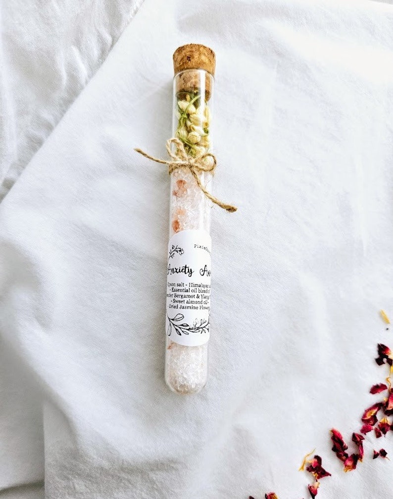 May include: A glass test tube filled with pink Himalayan salt, dried jasmine flowers, and a white label that reads "Anxiety Away" with a list of ingredients: Himalayan salt, essential oil blend of bergamot & ylang ylang, sweet almond oil, and dried jasmine flowers.