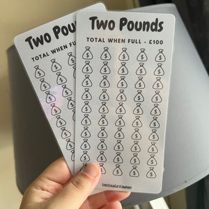 May include: Two white cards, each featuring the text "Two Pounds" and "TOTAL WHEN FULL - £100". The cards display a grid of 50 money bag icons. The cards are held in a hand.