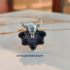 May include: A silver-toned ring featuring a bull skull design with curved horns and a cluster of black oval-shaped stones. The ring has the text "MOONLIGHTJEWELRYGIFT" at the bottom.