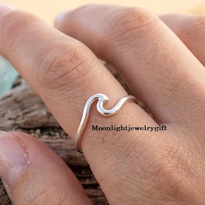 May include: A polished silver ring featuring a single continuous wave design that curves upward. The band is smooth and metallic, showcasing a minimalist aesthetic. The text Moonlightjewelrygift is visible on the band of the silver ring.