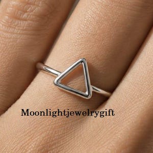 May include: A polished silver-toned ring featuring a simple, open triangle design on the band. The text Moonlightjewelrygift is visible in black lettering below the ring. The band is thin and smooth, showcasing a clean, geometric aesthetic.