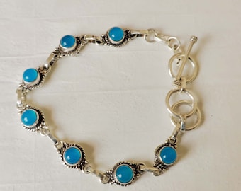 May include: A silver bracelet with a toggle clasp features six turquoise blue gemstone accents. The bracelet is made of a delicate chain with a simple design.