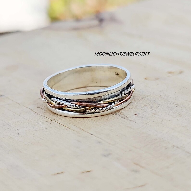 Pu&ograve; includere: Un anello d'argento con un designo intrecciato con accenti in rame e oro. L'anello &egrave; su una superficie di legno.