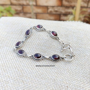 K&ouml;nnte beinhalten: Ein silbernes Armband mit roten Edelstein-Akzenten. Das Armband hat einen Karabinerverschluss.