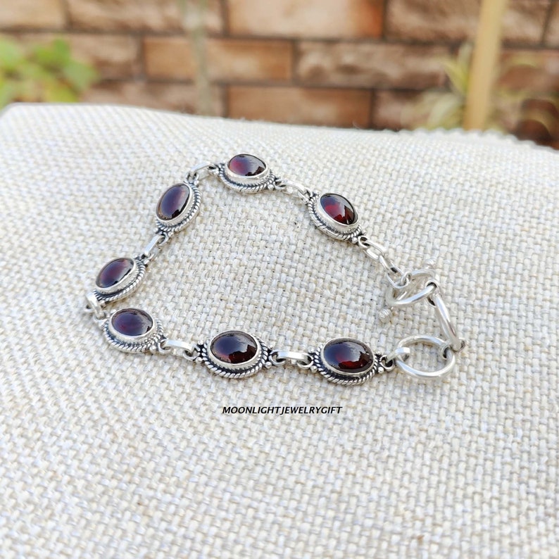 K&ouml;nnte beinhalten: Ein silbernes Armband mit roten Edelstein-Akzenten. Das Armband hat einen Karabinerverschluss.