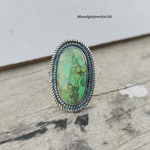 May include: Oval-shaped silver ring with a large, light green stone with gold-colored veining. The stone is set in a detailed silver bezel with a twisted rope design. The ring is displayed on a weathered wooden surface.