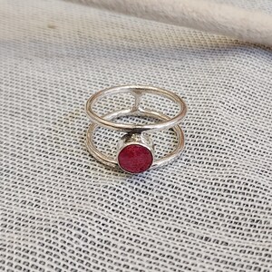 Może przedstawiać: Srebrny pierścionek z podw&oacute;jnym paskiem i czerwonym kamieniem szlachetnym w środku.