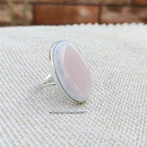 May include: A silver ring with a large, oval-shaped, light pink gemstone. The gemstone is surrounded by a silver band. The ring is set on a light-colored, textured surface. The text "MOONLIGHTJEWELRYGIFT" is visible.
