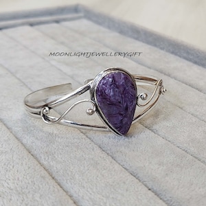 Pode incluir: Um bracelete de prata com uma pedra em forma de lágrima de cor púrpura e branca. A pedra está engastada em um bezel de prata e o bracelete tem um design delicado e em espiral.