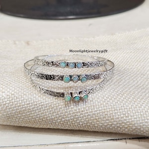May include: Three silver bangles with an ornate floral design and white opal stones. The bangles are stacked on top of each other on a beige fabric surface.