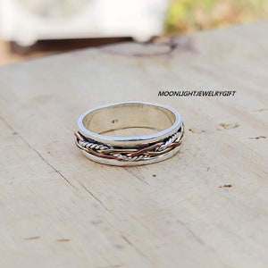 Pu&ograve; includere: Un anello a fascia d'argento con un design intrecciato in rame e filo d'argento. L'anello &egrave; su una superficie di legno.