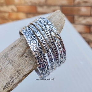 Pu&ograve; includere: Un set di sette braccialetti d'argento con intricati disegni floreali e geometrici. I braccialetti sono impilati su un pezzo di legno di deriva. Il testo "moonlightjewelrygift" &egrave; visibile nella parte inferiore dell'immagine.