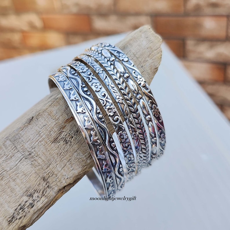 Pu&ograve; includere: Un set di sette braccialetti d'argento con intricati disegni floreali e geometrici. I braccialetti sono impilati su un pezzo di legno di deriva. Il testo "moonlightjewelrygift" &egrave; visibile nella parte inferiore dell'immagine.