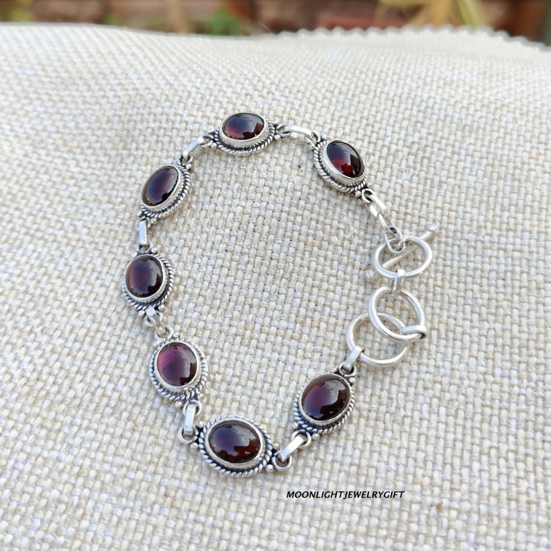 K&ouml;nnte beinhalten: Ein silbernes Armband mit einem Schiebeverschluss zeigt eine Reihe von ovalen, dunkelroten Edelsteinen. Das Armband liegt auf einem beigefarbenen Stoffhintergrund.