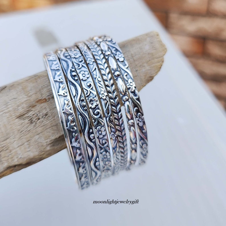 Pu&ograve; includere: Un set di nove braccialetti d'argento con diversi disegni in rilievo, tra cui motivi floreali, linee ondulate e trame intrecciate. I braccialetti sono impilati su un pezzo di legno galleggiante.