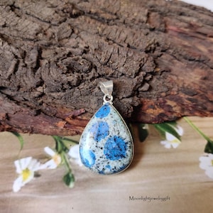Può includere: Ciondolo a goccia con anello argentato. Il ciondolo presenta una pietra blu e bianca con un motivo maculato, incastonata in una lunetta argentata. Lo sfondo è una superficie marrone strutturata con fiori bianchi.